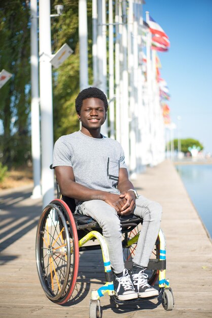 Retrato de hombre guapo en silla de ruedas. Hombre afroamericano con ropa informal en el terraplén, sonriendo. Cielo azul y banderas de fondo. Retrato, belleza, concepto de felicidad.