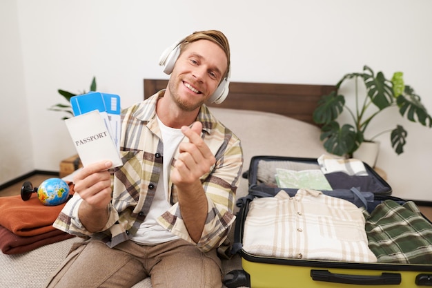 Foto gratuita retrato de un hombre guapo y sonriente con auriculares sentado con la maleta abierta en la cama muestra el pasaporte con