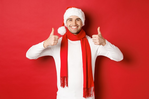 Retrato de hombre guapo sonriente con bufanda de santa y sombrero mostrando thumbsup celebrando el stand de navidad ...