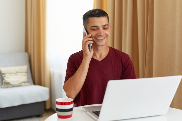 Retrato de hombre guapo sonriente feliz sentado frente a la computadora portátil blanca y hablando con alguien a través de un teléfono inteligente, hombre vestido con camiseta informal marrón, posando en casa en la sala de estar.