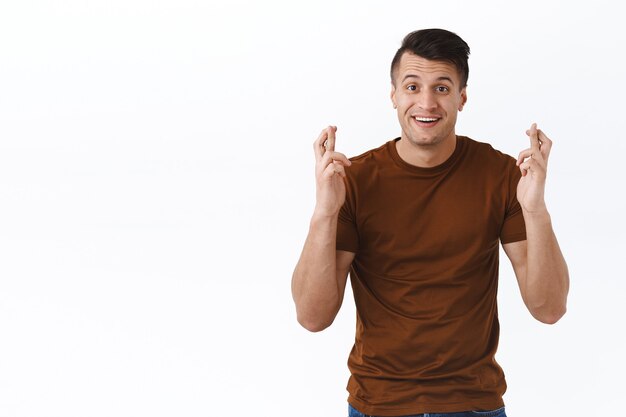 Retrato de hombre guapo sonriente y optimista rezando para que el sueño se haga realidad, anticipando buenas noticias, la fortuna de su lado, cruzar los dedos, buena suerte