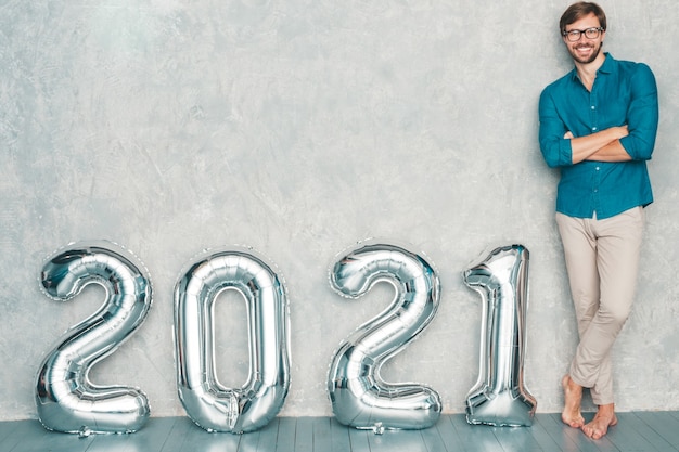 Retrato de hombre guapo sonriente posando junto a la pared. Hombre barbudo sexy staning cerca de Silver 2021 Balloons. Feliz año nuevo 2021. Números metálicos 2021