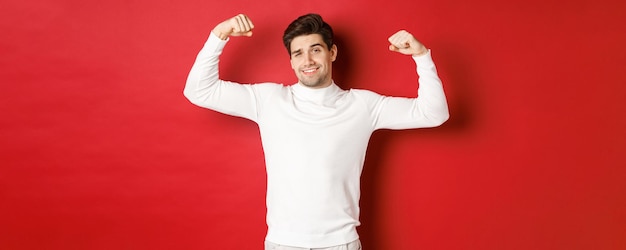Foto gratuita retrato de hombre guapo sonriente en suéter blanco flexionando bíceps y fanfarroneando con alarde de fuerza ...