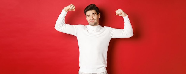 Retrato de hombre guapo sonriente en suéter blanco flexionando bíceps y fanfarroneando con alarde de fuerza ...