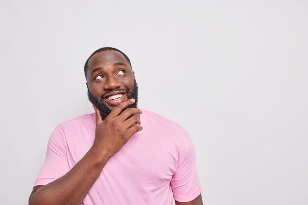 Retrato de hombre guapo con sonrisa con dientes sostiene la barbilla enfocada arriba vestido con camiseta rosa casual aislada sobre pared blanca