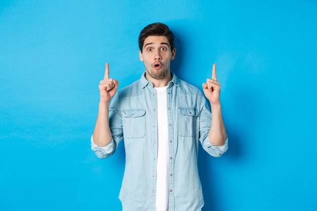 Retrato de hombre guapo sorprendido apuntando con el dedo hacia arriba, mostrando el logo sobre fondo azul.