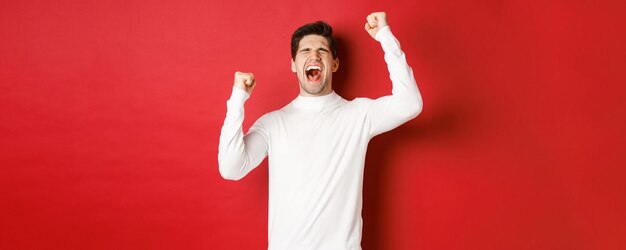 Retrato de un hombre guapo con suéter blanco, sintiéndose alegre, celebrando la victoria, gritando de alegría y levantando las manos en victoria, de pie sobre un fondo rojo.