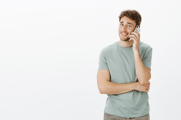 Retrato de hombre guapo tímido con cerdas, hablando por teléfono inteligente y sonriendo ampliamente