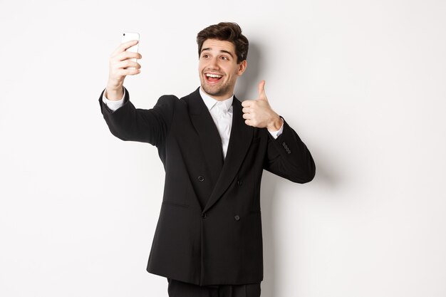 Retrato de hombre guapo tomando selfie en fiesta de año nuevo, vestido con traje, tomando fotos en el teléfono inteligente y mostrando el pulgar hacia arriba, de pie contra el fondo blanco.