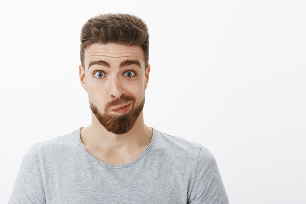 Retrato de hombre guapo tonto y divertido con barba, bigote y ojos azules sonriendo burlonamente haciendo cara incómoda e incómoda mirando en el espejo y pensando en hacer cambios posando contra la pared gris