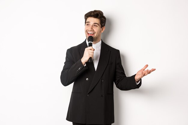 Retrato de hombre guapo en traje negro cantando una canción, sosteniendo el micrófono y dando un discurso, de pie contra el fondo blanco.