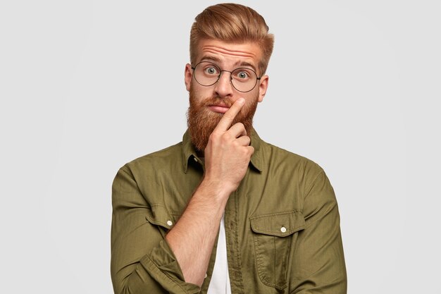 Retrato de hombre guapo vacilante con espesa barba pelirroja, se ve sorprendentemente, se pregunta las últimas noticias, vestido con ropa de moda, aislado sobre una pared blanca. Expresiones faciales