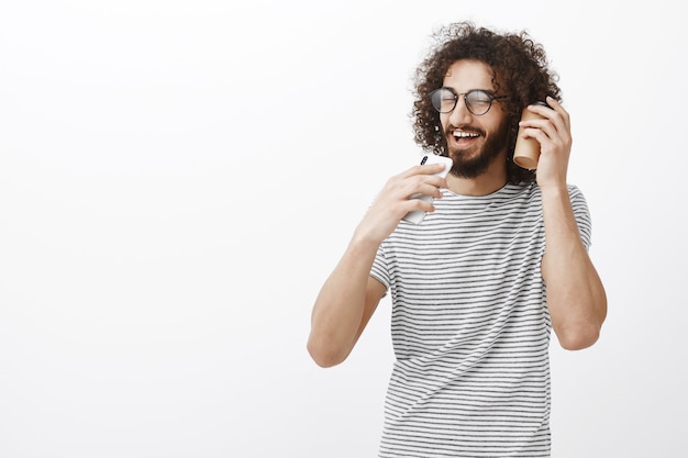 Retrato de hombre guapo yute despreocupado con barba en gafas negras, sosteniendo un teléfono inteligente como micrófono y cantando junto con una taza de café cerca de la oreja