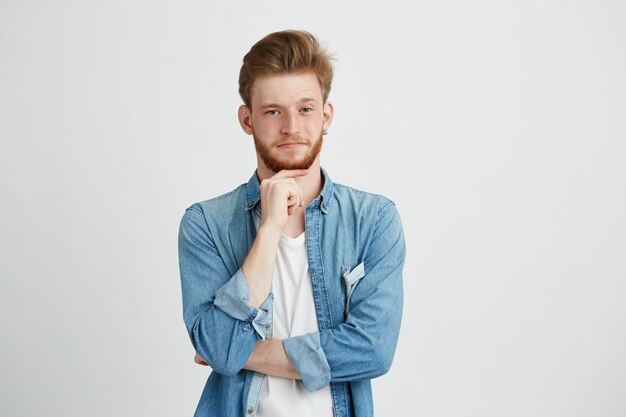 Retrato del hombre hermoso joven confiado con la barba que piensa con la mano en la barbilla.