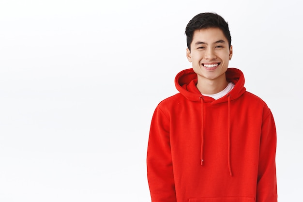 Retrato de hombre hipster asiático feliz y sonriente, chico joven con sudadera con capucha roja sonriendo alegre, mirando la cámara entusiasta, expresando un estado de ánimo positivo, estar encantado o satisfecho, pared blanca.