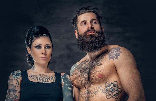 Retrato de hombre hipster barbudo sin camisa y tatuado y mujer morena con tinta de tatuaje en el torso sobre fondo gris oscuro.