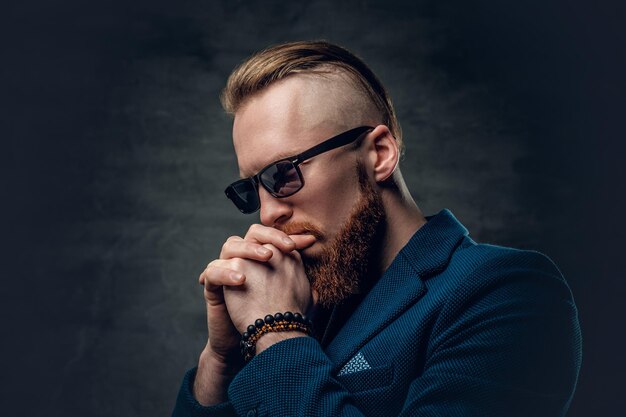 Retrato de hombre hipster barbudo pelirrojo vestido con una chaqueta azul y gafas de sol sobre fondo gris.