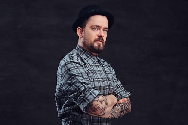 Retrato de un hombre hipster tatuado de mediana edad con barba y peinado vestido con una camisa a cuadros y un sombrero, posa con los brazos cruzados en un estudio. Aislado en un fondo de textura oscura.