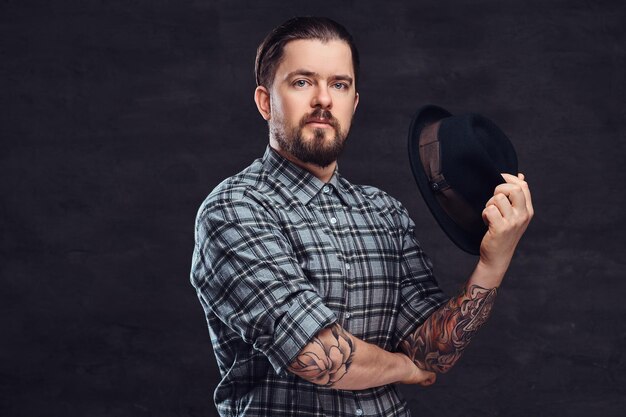 Retrato de un hombre hipster tatuado de mediana edad con barba y peinado vestido con una pose de camisa a cuadros con un sombrero en la mano. Aislado en un fondo de textura oscura.