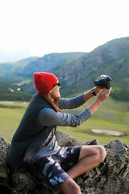 Retrato de hombre hipster viajando a las montañas, usa sombrero rojo y ropa hipster, toma fotos