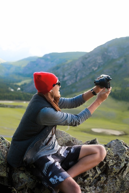 Retrato de hombre hipster viajando a las montañas, usa sombrero rojo y ropa hipster, toma fotos