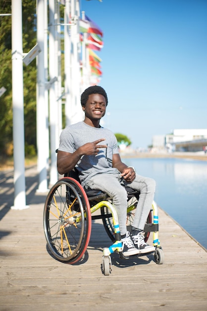 Retrato de un hombre indiferente en silla de ruedas. Hombre afroamericano con ropa informal en el terraplén, mostrando el signo de la victoria. Cielo azul y banderas de fondo. Retrato, belleza, concepto de felicidad.