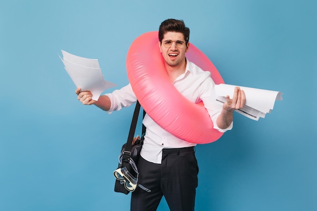 Retrato de hombre insatisfecho con muchos documentos Chico con gafas y traje de oficina posando con círculo inflable sobre fondo azul