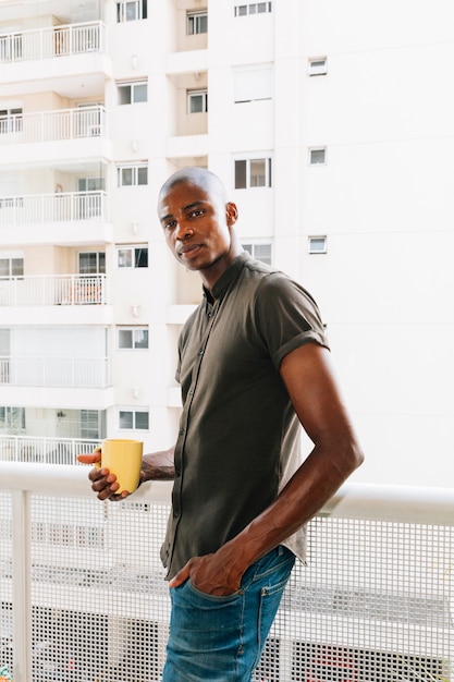 Retrato de un hombre joven afro que se coloca en el balcón que sostiene la taza de café amarilla disponible
