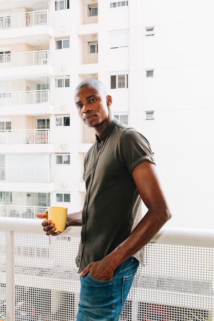 Retrato de un hombre joven afro que se coloca en el balcón que sostiene la taza de café amarilla disponible