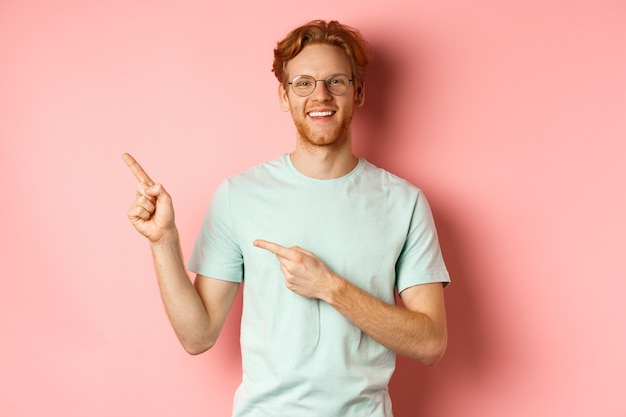 Retrato de hombre joven alegre con pelo rojo con gafas señalando con el dedo en la esquina superior izquierda ...