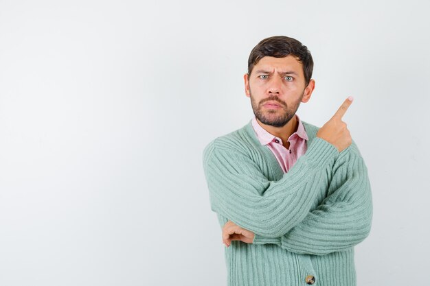 Retrato de hombre joven apuntando a la esquina superior derecha en camisa, chaqueta de punto y mirando vacilante vista frontal