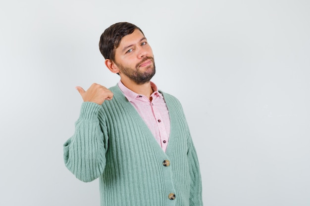 Retrato de hombre joven apuntando con el pulgar hacia atrás en camisa, chaqueta de punto y mirando confiado vista frontal