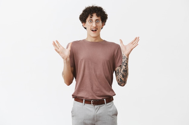 Retrato de hombre joven asombrado, atractivo, elegante y artístico con bigote, pelo rizado y brazo tatuado, gesticulando con las palmas levantadas, dando forma a un gran objeto, impresionado y asombrado con el tamaño