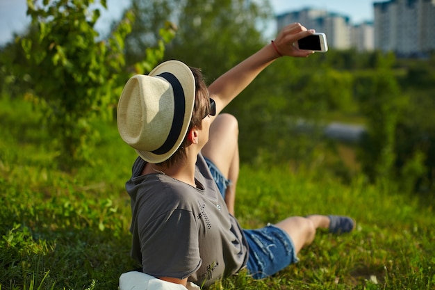 Retrato de hombre joven atractivo moderno con estilo en ropa casual con sombrero en gafas sentado en el parque en la hierba verde haciendo selfie