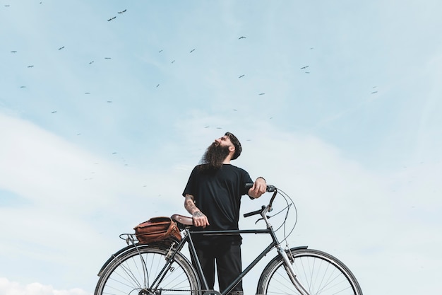 Foto gratuita retrato de un hombre joven con una bolsa en su bicicleta mirando pájaros volando en el cielo