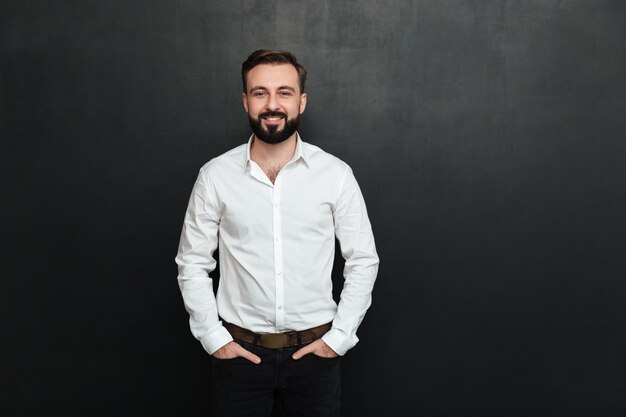 Retrato de hombre joven en camisa blanca posando en la cámara con una amplia sonrisa, y las manos en los bolsillos sobre gris oscuro