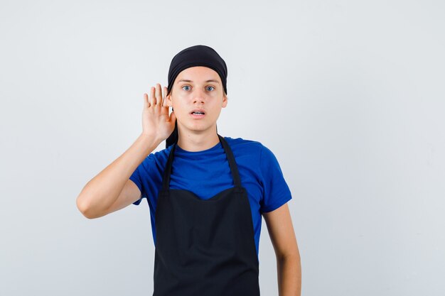Retrato de hombre joven cocinero con la mano detrás de la oreja en camiseta, delantal y mirando seria vista frontal