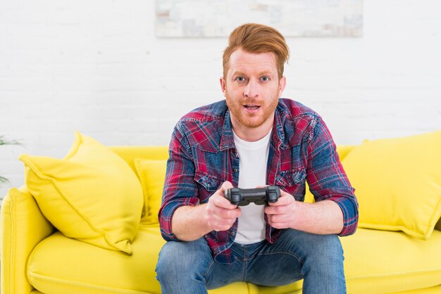 Retrato de un hombre joven emocionado que se sienta en el sofá amarillo que juega al videojuego