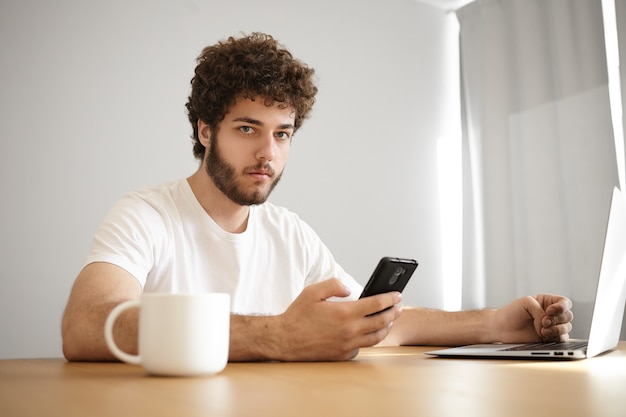 Retrato de hombre joven con estilo serio con rastrojo sosteniendo teléfono móvil marcando a su amigo mientras navega por internet en una computadora portátil genérica, tomando una bebida caliente en la mesa de madera en el interior,