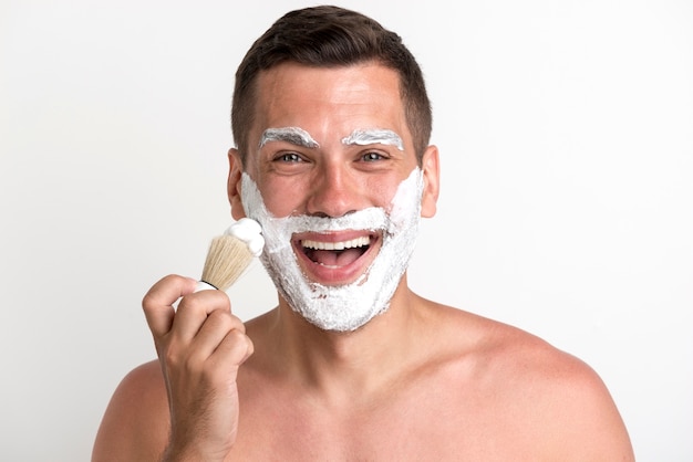 Retrato de hombre joven feliz aplicando espuma de afeitar sobre fondo blanco.