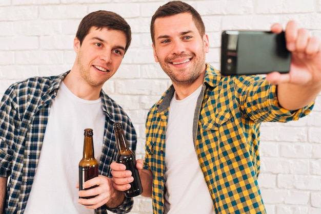 Foto gratuita retrato del hombre joven feliz que sostiene la botella de cerveza disponible que toma el selfie con sus amigos en smartphone