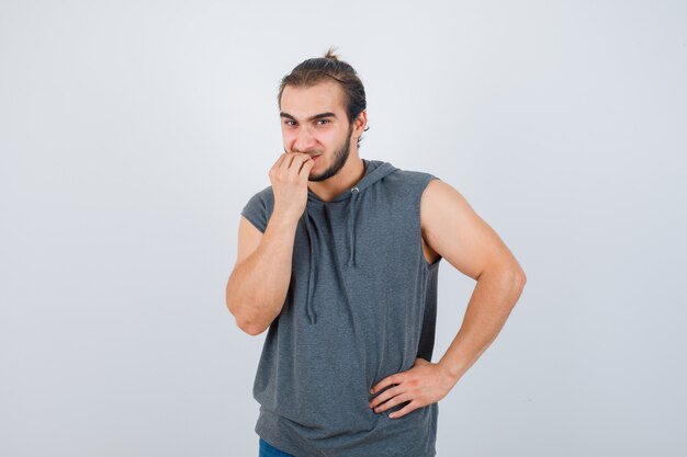 Retrato de hombre joven en forma mordiendo las uñas en sudadera con capucha sin mangas y mirando estresado vista frontal