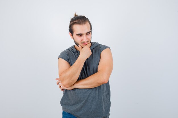 Retrato de hombre joven en forma de pie en pose de pensamiento en sudadera con capucha sin mangas y mirando pensativo vista frontal