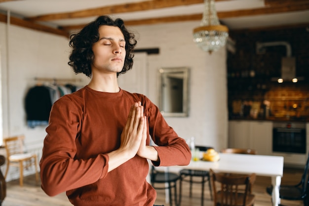 Retrato de hombre joven guapo con cabello rizado negro posando en el acogedor interior de la cocina manteniendo los ojos cerrados y presionando las manos juntas en namaste, rezando, haciendo meditación, con mirada tranquila