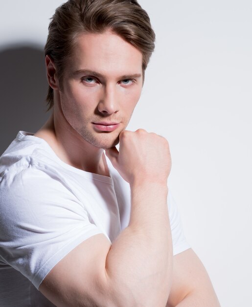 Retrato de hombre joven guapo con la mano cerca de la cara en camiseta blanca posa sobre la pared con sombras de contraste.