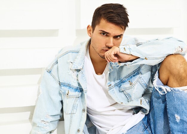 Retrato de hombre joven y guapo modelo vestido con ropa de jeans sentado cerca de la pared blanca con textura