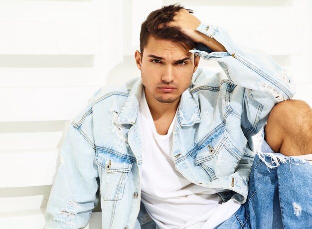Retrato de hombre joven y guapo modelo vestido con ropa de jeans sentado cerca de la pared blanca con textura