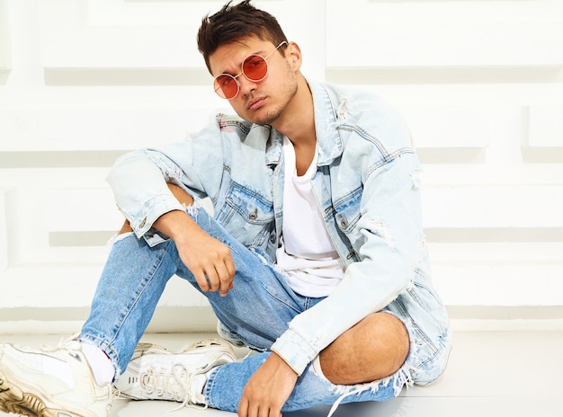 Retrato de hombre joven y guapo modelo vestido con ropa de jeans sentado cerca de la pared blanca con textura