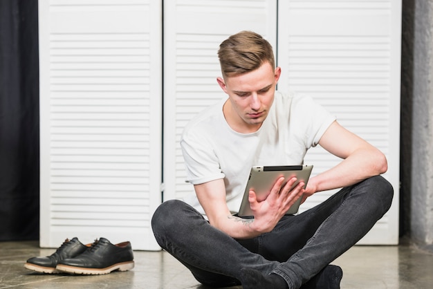 Retrato de hombre joven guapo sentado en el piso usando tableta digital en la mano