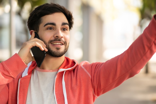 Retrato de hombre joven hablando por teléfono y levantando la mano para llamar a un taxi mientras está parado al aire libre en la calle. Concepto urbano.